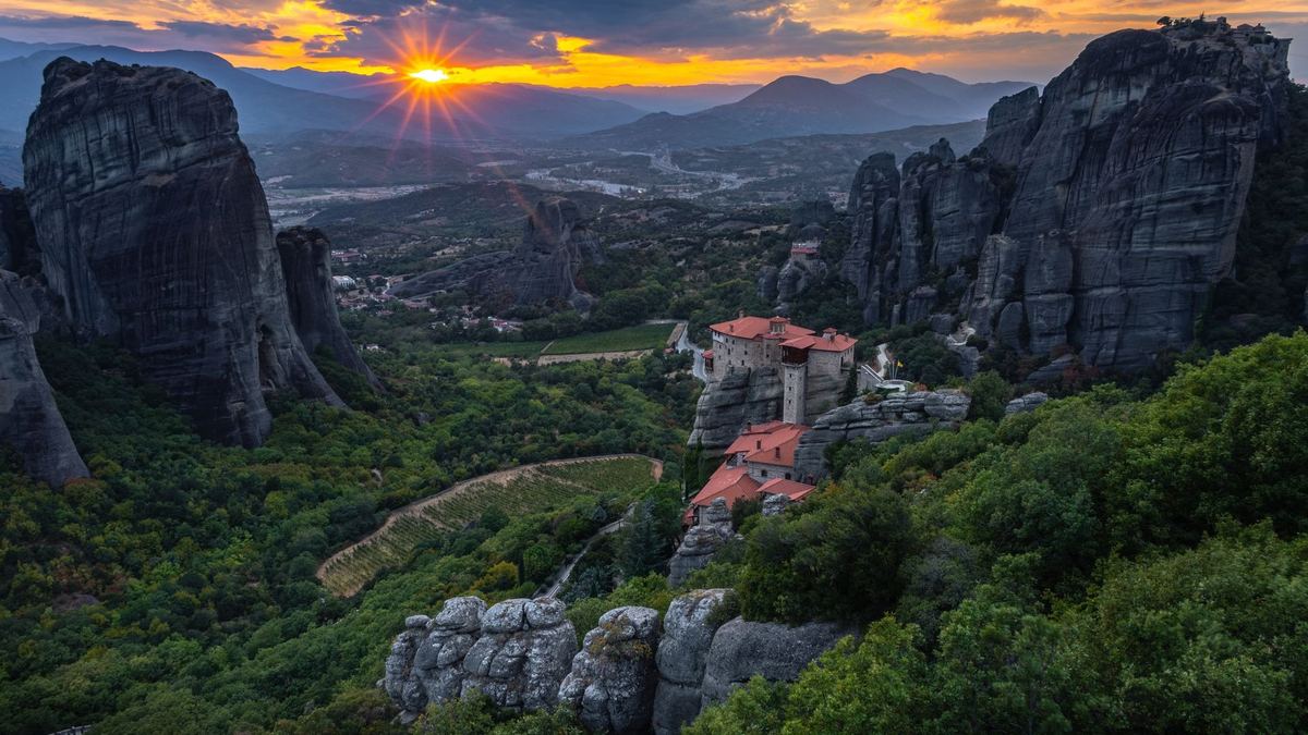 The sun setting behind the Meteora rock towers and a red-roofed monastery